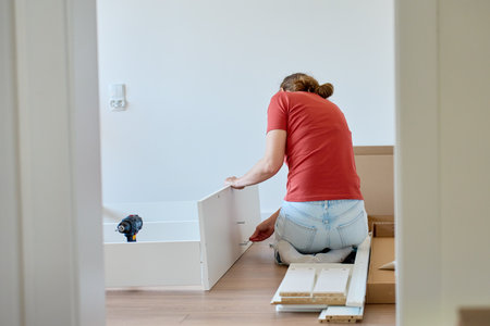 Woman sits on wooden floor and assembling furniture in new home, fastens parts of white bed frame. DIY home renovation and home organization after moving conceptの写真素材