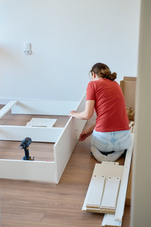 Woman sits on wooden floor and assembling furniture in new home, fastens parts of white bed frame. DIY home renovation and home organization after moving conceptの写真素材