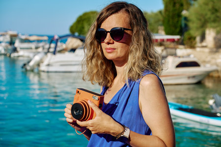 Woman photographing with instant camera near marina with boats in background. Female tourist taking photo by sea water with retro camera Summer vacation and travel conceptの写真素材