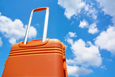 Bright orange suitcase with extended handle stands against blue sky with white clouds. Concept of travel, vacation and summer adventureの写真素材