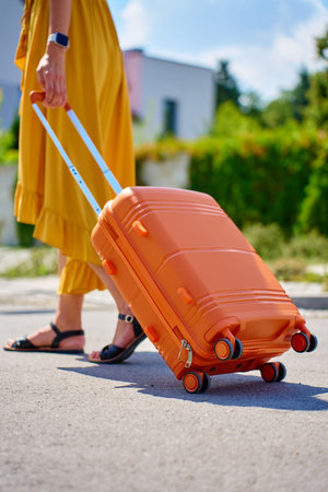 Woman in yellow dress with orange suitcase walking on street near residential house on sunny day. Female traveler with luggage return home from journeyの写真素材