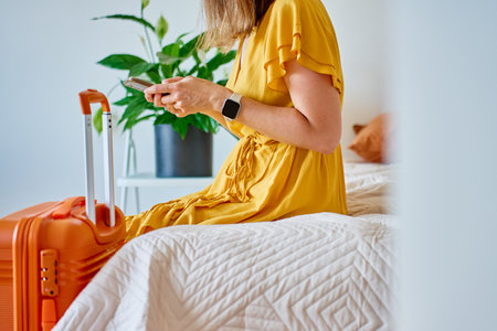 Woman in yellow dress sitting on bed with orange suitcase and using smartphone. Female traveler waiting for departure in modern interior with green houseplant. Concept of travel and trip preparationの写真素材