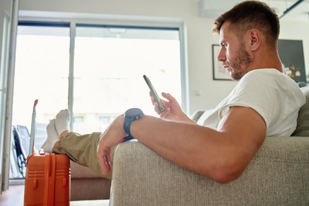Man sitting on sofa near orange suitcase and using smartphone, waiting for leaving home. Male traveler checking information in mobile phone before trip. Travel planning conceptの写真素材
