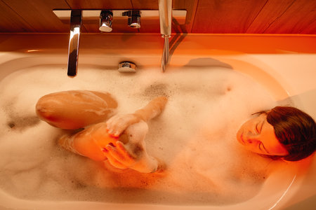 Woman lying in bathtub filled with foam and washing her legs. Female relaxing in bathroom with candles and ambient lighting Concept of wellness, relaxation, and self careの写真素材