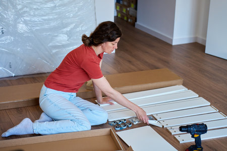 Woman unpacking parts for furniture assembly at home. Female sitting on floor and holding bed parts, wheels and hardware. Concept of DIY home improvement and renovationの写真素材