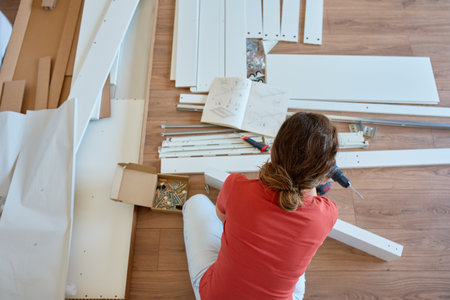 Woman assembling white furniture parts on wooden floor using drill, work tools and instruction...