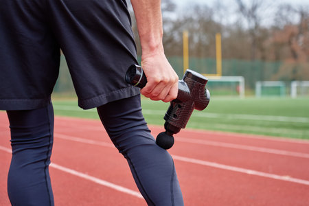 Close-up of man recovery muscle after training. Athlete using massage gun on leg while standing on running track. Concept of muscle recovery, sports therapy and injury preventionの写真素材