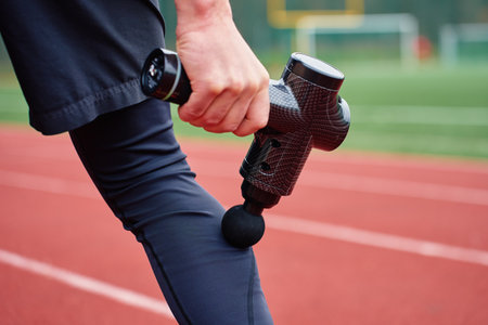 Close-up of man recovery muscle after training. Athlete using massage gun on leg while standing on running track. Concept of muscle recovery, sports therapy and injury preventionの写真素材