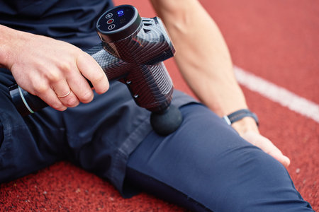 Close-up of man sitting on red running track using black massage gun on his leg after training. Concept of muscle recovery, sports therapy, and injury prevention for active lifestylesの写真素材