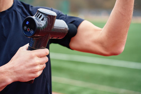 Close-up of man in black shirt using massage gun on his arm muscle at outdoor sports field after training. Concept of muscle recovery, sports therapy and injury preventionの写真素材