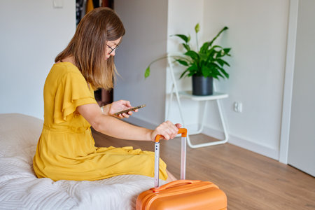 Woman in yellow dress sitting on bed near orange suitcase and using smartphone, waiting for leaving home. Female traveler checking information in mobile phone before trip. Travel planning conceptの写真素材