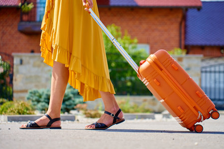 Woman in yellow dress with orange suitcase walking on street near residential house on sunny day. Female traveler with luggage return home from journeyの写真素材