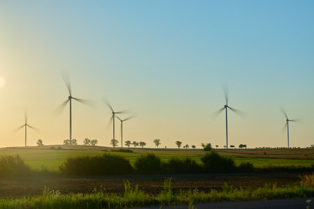 Wind turbines in farmland with green fields and trees under evening sky. Windmills for production clean energy at sunset. Concept of renewable energy, sustainable resources and green technologyの写真素材
