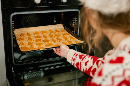 Woman in red festive sweater and Santa hat putting tray of Christmas cookies into oven. Process of cooking traditional gingerbread cookies for winter holidays at home kitchenの写真素材