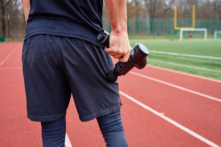 Close-up of man recovery muscle after training. Athlete using massage gun on leg while standing on running track. Concept of muscle recovery, sports therapy and injury preventionの写真素材