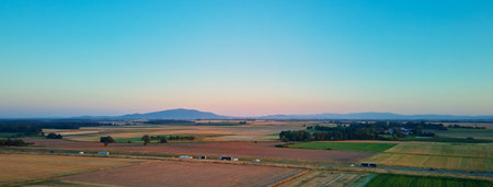 Aerial view of countryside landscape with farmland, highway road with cars among fields and mountain range in background at sunsetの写真素材