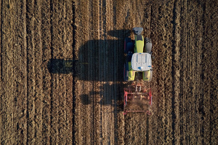 Green tractor with machinery working on agricultural field, plowing farmland under clear sky, aerial view. Small farming conceptの写真素材