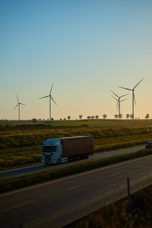 Vehicles driving on highway with wind turbines on fields in background at sunset. Concept of renewable energy, transport and sustainabilityの写真素材