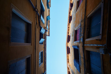 Upward view of narrow alley in Venice with open wooden shutters and clear blue sky between old buildings. Concept of urban architecture, history and European travelの写真素材