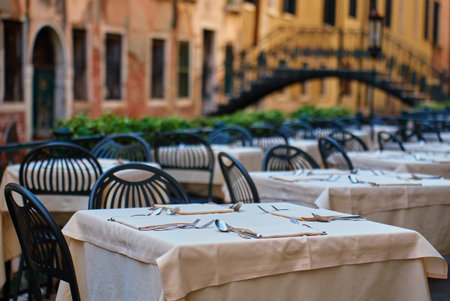 Empty outdoor restaurant tables with tablecloths and cutlery in Venice, Italy. Street cafe with bridge over Venetian canal on background in morning. Concept of dining, tourism and travelの写真素材