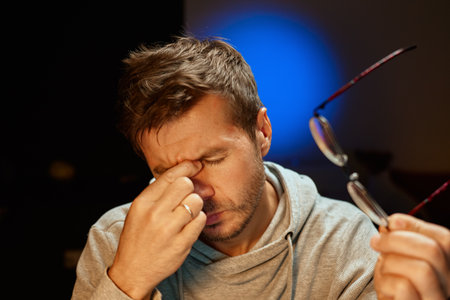 Tired man rubs his eyes while holding his glasses in hand. Fatigued person working late. Concept of eye strain, stress, headache and overworkの写真素材