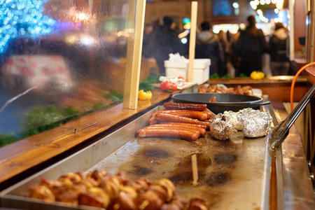 Close-up of grilled sausages and baked potatoes wrapped in foil on hot grill at street food stall during holiday fair. Process of cooking street food on Christmas marketの写真素材
