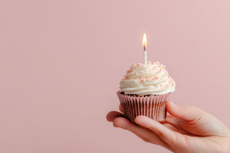Cupcake with white cream and lit candle in hand on pink background with copy space. Concept of birthday celebration, anniversary and festive dessertの素材