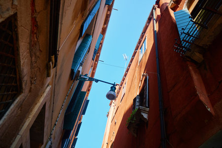 Upward view of narrow alley in Venice with open wooden shutters and clear blue sky between old buildings. Concept of urban architecture, history and European travelの写真素材