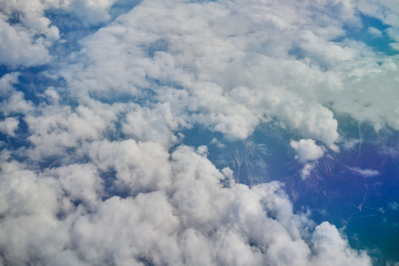 Aerial view from airplane window of white fluffy clouds floating above landscape, with mountains and riversの写真素材