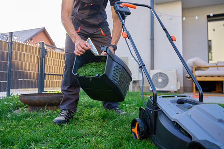 Person emptying grass container of electric lawn mower after grass moving. Man in work overalls working in backyard of suburban house. Concept of gardening, yard care and home maintenanceの写真素材