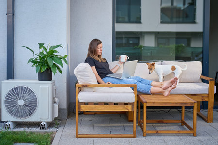 Woman and her dog sitting on terrace sofa with laptop and coffee cup. Concept of remote work, relaxation and life with pet at homeの写真素材