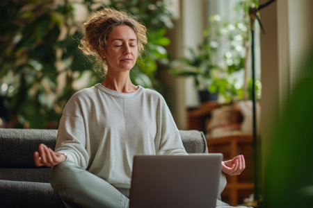 Young woman sitting cross-legged with closed eyes meditating in front of laptop at home. Concept of mindfulness, yoga and stress reliefの素材
