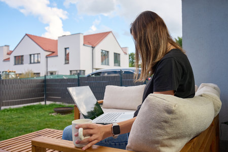 Woman sitting on sofa using laptop with wireless mouse and coffee cup. Person relaxing on home patio with wooden outdoor furniture. Concept of remote work, comfort and modern home lifestyleの写真素材