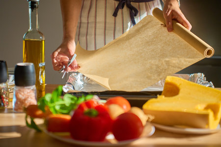 Woman in apron placing baking paper on tray in sunlight kitchen with fresh vegetables, pumpkin, species and olive oil on table. Concept of home cooking, preparation and healthy homemade foodの写真素材