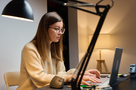 Woman working at desk with laptop in home office, sitting at table with lamp and looking at screen. Remote work at nightの写真素材