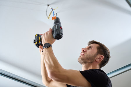 Man wearing work overalls drills hole in ceiling using cordless power drill in indoor room. Concept of home renovation, installation and professional maintenance service.の写真素材