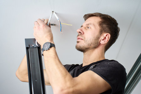 Man in workwear connecting electrical wires while installing ceiling light fixture indoors. Concept of lighting installation, electrical work and home interior repairの写真素材