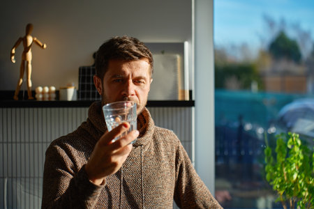 Adult man drinking fresh clean water from glass while standing in kitchen near window with sunlight....