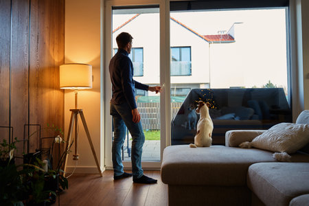 Man standing near large window in living room and looking outside while dog sits on sofa. Concept of...