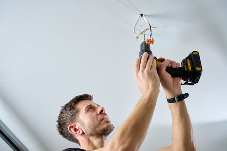 Man wearing work overalls drills hole in ceiling using cordless power drill in indoor room. Concept of home renovation, installation and professional maintenance service.の写真素材