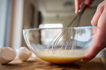 Hands whisk beaten eggs in glass bowl on wooden kitchen counter with cracked eggshells. Person preparing breakfast. Concept of home cooking, food preparation and recipe ingredients.の写真素材