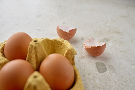 Brown chicken eggs in paper carton and two cracked empty eggshell halves on light kitchen...