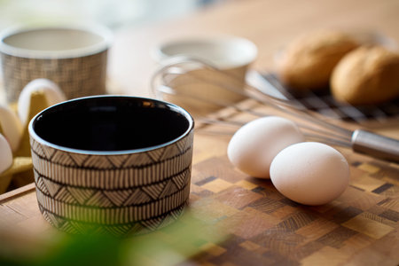 Two whole eggs on cutting board near patterned ceramic bowl and metal whisk on wooden cutting board...