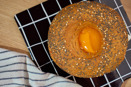 Top view of seeded bagel with baked egg yolk on black grid cutting board with gold cutlery and...