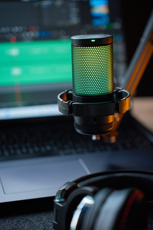 Illuminated studio microphone on boom arm on desk with headphones and laptop with audio editing...