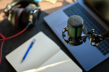 Close up of podcast microphone above laptop, headphones, and notebook on desk. Concept of recording,...