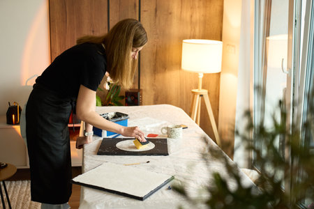 Woman makes handmade wall decor on black canvas, pressing a sponge on a white plaster circle at a covered table. Concept of DIY craft and textured art.