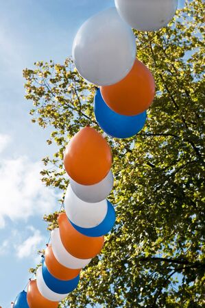 Blue, white and orange balloons hanging in line in front of a blue sky and a tree. Focus on the balloons in the middle.の写真素材