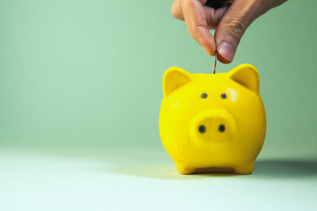Hand putting coin into yellow piggy bank on green background with copy space