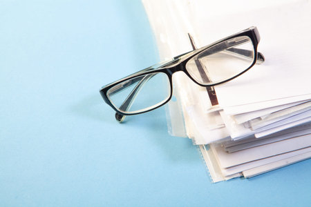 Pile of papers and eyeglasses on blue background with copy space
